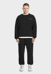 Czarny zestaw bluz i spodni dresowych, z luźnym krojem, prążkowanymi mankietami i białą wewnętrzną warstwą. Buty są białe z kolorowymi akcentami.