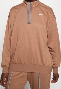 Brun, texturerad tröja med hög krage och dragkedja, med en svart och vit mönstrad detalj längs dragkedjan. Nike-logotyp på bröstet.