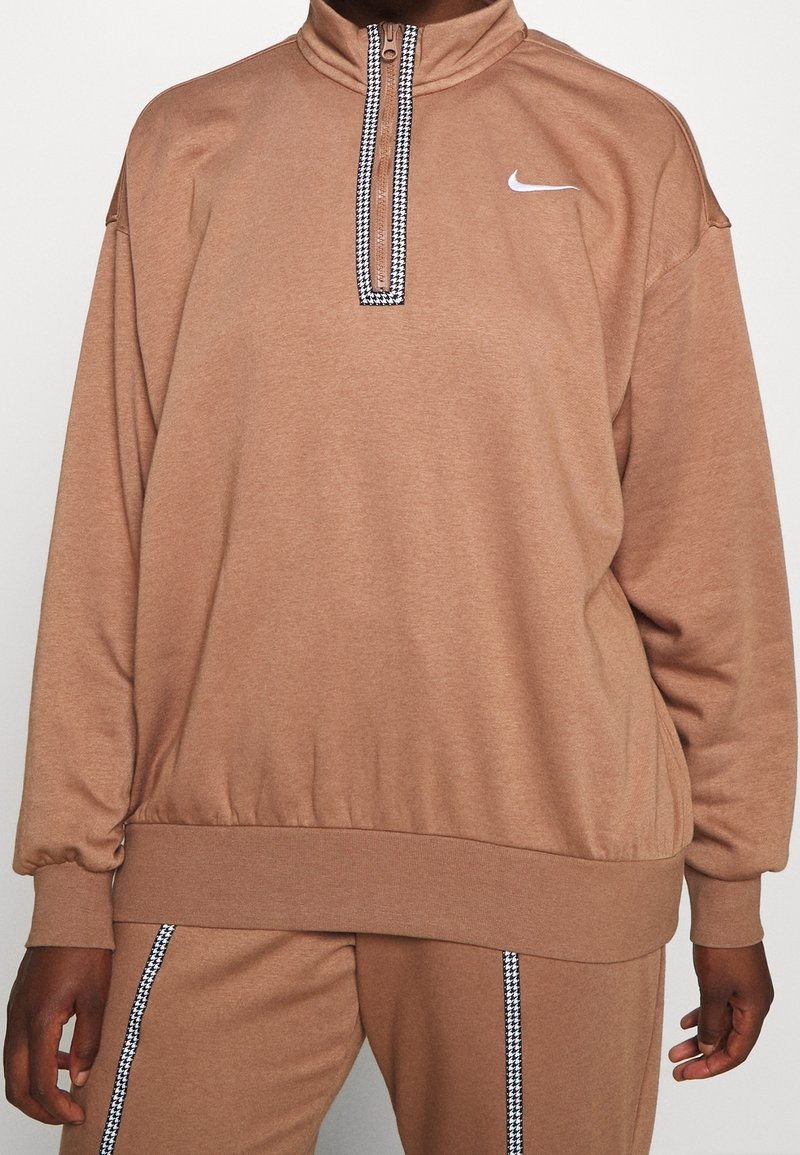Brun, texturerad tröja med hög krage och dragkedja, med en svart och vit mönstrad detalj längs dragkedjan. Nike-logotyp på bröstet.