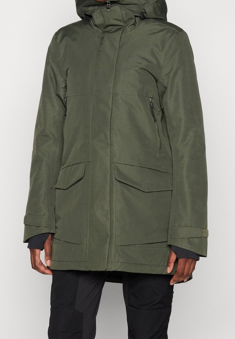 Homme portant une parka à capuche vert olive avec de grandes poches avant et un pantalon noir, debout devant un fond uni.