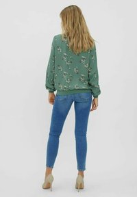 Kobieta z długimi blond włosami, ubrana w zieloną kwiecistą bluzkę, niebieskie dopasowane jeansy i beżowe buty na wysokim obcasie, stoi tyłem na jednolitym tle.