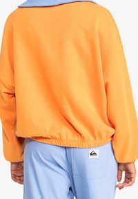 Felpa arancione oversize con colletto blu; orlo elasticizzato in vita. Pantaloni azzurri chiari con tasca posteriore e patch con logo.