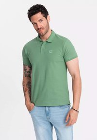 Zelená polo košile vyrobená z hladkého bavlněného materiálu, s malým vyšitým logem na hrudi, krátkými rukávy a klasickým límečkem.