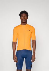 Maillot de cyclisme à manches courtes en orange vif avec une fermeture éclair à l'avant et un logo noir ; associé à un short de cyclisme bleu marine portant un logo.