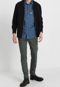 Černá zipová bunda přes denímovou košili, zelené slim-fit kalhoty a černé kotníkové boty. Outfit se vyznačuje klasickými liniemi a neformálním stylem.