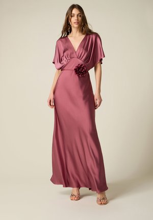 Donna che indossa un abito lungo in raso color malva con scollo a V profondo, maniche svolazzanti, dettaglio floreale in vita e sandali con tacchi alti e cinturini.