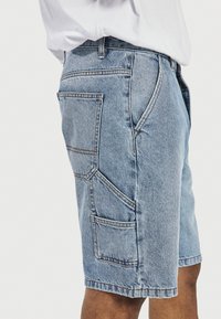 Hele sinised teksast lahtised lühikesed püksid mitme taskuga, mida kannab valget särki kandev inimene, näidatud külje pealt vööst põlvedeni.