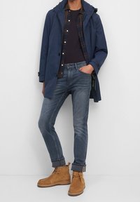 Námořnický voděodolný kabát přes kostkovanou košili, v kombinaci s tmavě modrými džíny a světlými semišovými botami. Mezi vlastnostmi je kapuce a ohrnuté manžety.