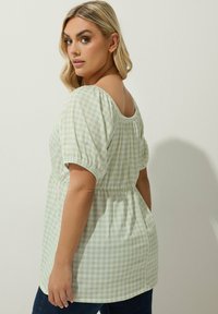 Blus dengan pola gingham hijau dan putih, berlengan pendek puff, dengan leher lebar dan pinggang yang dikecilkan, dipadukan dengan celana jeans gelap.