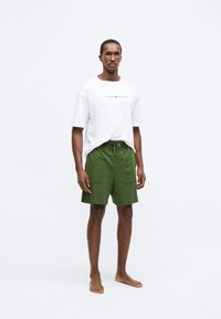 Λευκό μπλουζάκι με κόκκινο λογότυπο και πράσινο σορτσάκι. Το σορτσάκι διαθέτει ελαστική μέση με κορδόνι. Το μοντέλο στέκεται ξυπόλυτο σε ουδέτερο φόντο.