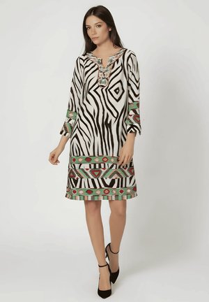 Mujer con vestido blanco y negro a rayas de cebra con patrones geométricos en verde, rojo y beige, que lleva zapatos de tacón negros con correa en el tobillo, de pie sobre un fondo blanco.