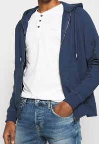 Granatowa bluza z kapturem na zamek z przednimi kieszeniami, wykonana z mieszanki bawełny, z tonalnym logo na rękawie. Pod spodem biała koszulka henley z trzema guzikami.