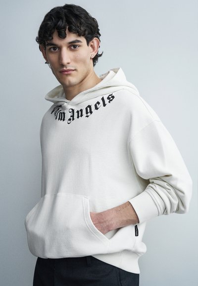 Jeune homme aux cheveux bouclés et foncés portant un sweat à capuche blanc avec le texte « Palm Angels », les mains dans la poche avant, debout devant un arrière-plan neutre.