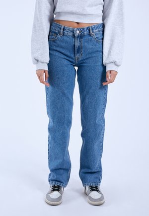 Persoon draagt lichtblauwe hoogwaterjeans, grijze cropped sweater en wit-grijze sneakers, staand tegen een effen witte achtergrond.