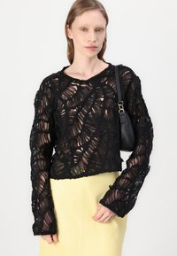 Schwarzer, transparenter, strukturierter Pullover mit einem Blättermuster, ausgestattet mit langen Ärmeln und einem runden Ausschnitt, kombiniert mit einem gelben Rock und einer schwarzen Tasche.