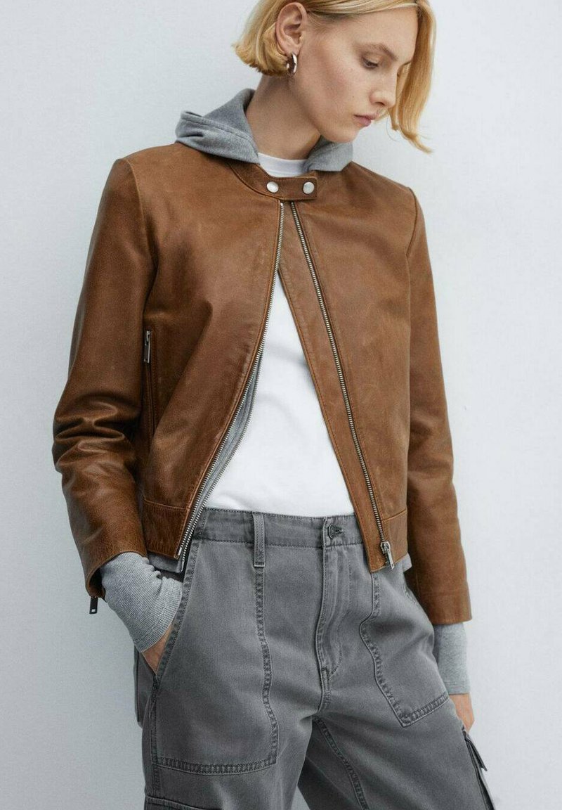 Brązowa skórzana kurtka z szarym kapturem, z zamkiem z przodu, zapięciem na napy i bocznymi kieszeniami, w połączeniu z szarymi spodniami cargo.