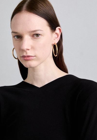 Top negro de cuello en v, con una textura suave. Pendientes de aro dorados realzan el look, en un fondo neutro. La piel parece natural.