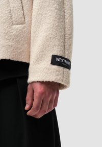 Kurtka polarowa w kolorze kremowym o strukturalnym designie, z czarną metką na mankiecie z napisem "WRSTBHVR STUDIO". Teksturowana powierzchnia.