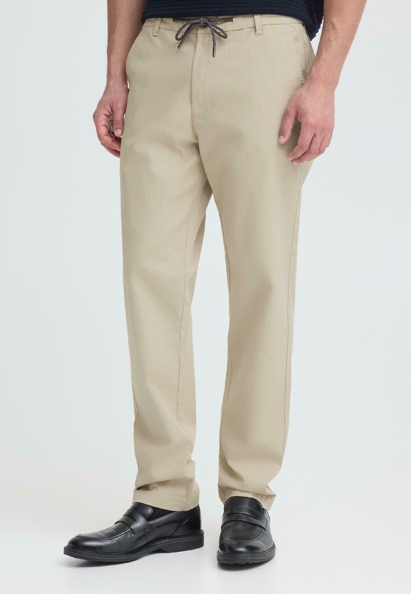 Homme portant un pantalon beige à cordon de serrage et des chaussures en cuir noires à enfiler, debout devant un fond clair.