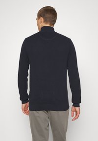 Navy knit pullover with a high collar, long sleeves, and ribbed cuffs. Textured fabric shows a subtle, woven pattern.