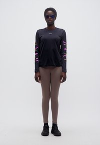 Άτομο που φοράει μαύρο μακρυμάνικο μπλουζάκι με μοτίβα σε μωβ και κίτρινο στα μανίκια, κολάν σε απόχρωση ταυπ, μαύρα αθλητικά παπούτσια και μπλε γυαλιά ηλίου με αντανακλαστικό φακό, σε λευκό φόντο.