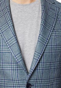 Zielono-niebieska marynarka w kratę o teksturowanym splocie, z klapą w kształcie notcha i jednym guzikiem. Nosi ją na lekko szarym t-shircie.