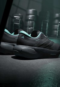 Paire de chaussures de course noires et turquoise étiquetées "Supernova Rise 3" et "Prime X", posées sur un sol sombre avec des pneus empilés reflétés en arrière-plan.