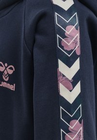 Granatowy sweter z różowym logo i wzorzystą boczną wstawką z kwiatowymi wzorami oraz kształtami zygzakowymi w kontrastujących kolorach.