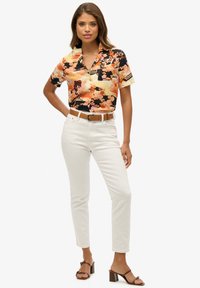 Camisa de manga corta con estampado floral en negro, naranja y crema, con cuello y bolsillo en el pecho, combinada con pantalones blancos ajustados y sandalias marrones de tacón.