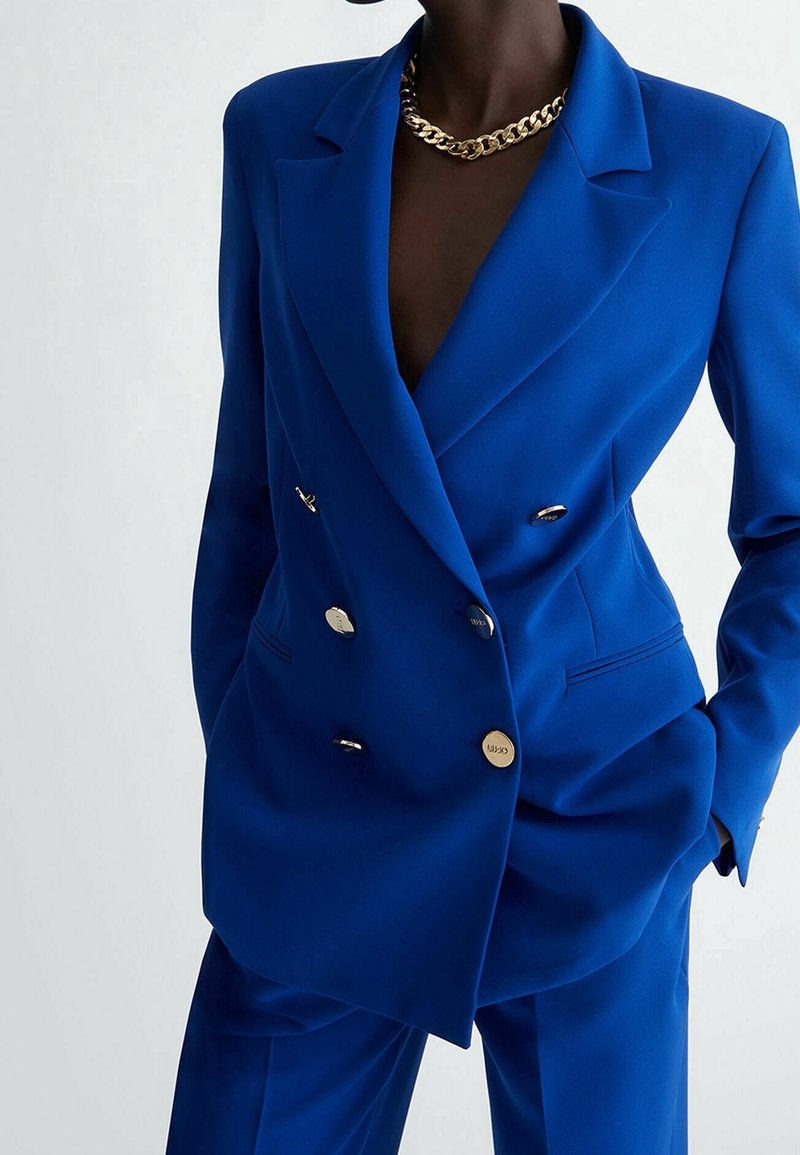 Niebieska marynarka dwurzędowa o dopasowanym kroju, z srebrnymi guzikami, ostrymi klapami i gładką fakturą tkaniny.