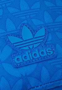 Blå Adidas patch med en blomlogotyp som har tre blad. Den texturerade bakgrunden visar ett subtilt mönster, kompletterat med stickade detaljer.