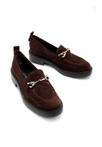 Brązowe zamszowe loafersy z uniesioną częścią palcową, dekoracyjnym srebrnym zdobieniem i grubą czarną gumową podeszwą. Podwójne uchwyty na pięcie.