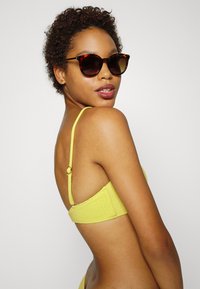 Top de bikini amarelo texturizado com tiras finas e fecho nas costas, usado com óculos de sol com armação tartaruga por uma modelo com cabelo curto e encaracolado.