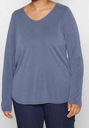 Persoon draagt een langermelig shirt met ronde hals in een gedempte blauwe kleur en donkere blauwe spijkerbroek, staand tegen een eenvoudige lichte achtergrond.