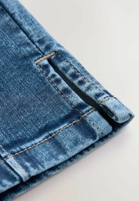 Cuffs od trapera s svijetlim plavim pranjem, s vidljivim šavovima, malim bočnim prorezom i glatkom teksturom.