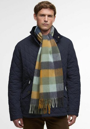 Geruite wollen sjaal met groene, blauwe, gele en grijze patronen en franjeranden, gedragen over een marineblauwe doorgestikte jas.