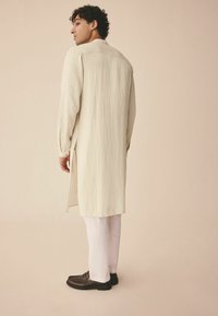 Langer, cremefarbener, strukturierter Mantel mit seitlichen Schlitzen, kombiniert mit weißen Hosen und dunkelbraunen Schuhen. Einfaches und strukturiertes Design.