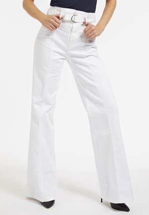 Vrouw draagt witte, wijde broek met hoge taille, bijpassende riem en zwarte puntige hakken, handen die de riemlussen aanpassen.