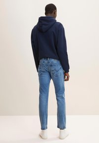 Hanorac bleumarin cu șnur, blugi albaștri din denim cu un finisaj pălit și adidași albi; ținută casual prezentată din spate.