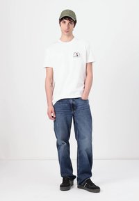 White cotton T-shirt featuring a small graphic, paired with loose blue denim jeans and black sneakers. The cap is olive green.