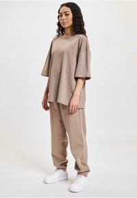 Overdimensjonert beige bomullst-skjorte med korte ermer, kombinert med matchende løse bukser. Hvite sneakers fullfører antrekket og tilfører kontrast.