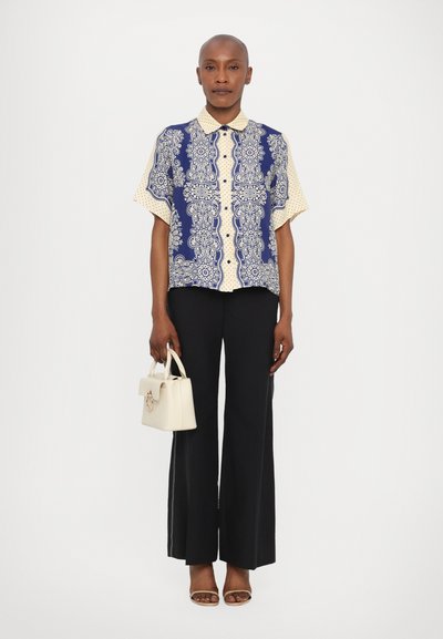 Camicia a maniche corte blu navy e beige con motivo, indossata con pantaloni neri a vita alta e con una piccola borsa bianca.