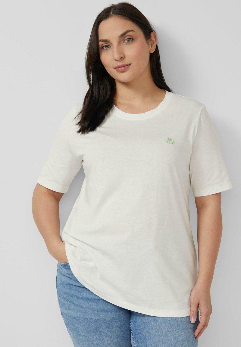 Femme aux cheveux bruns portant un t-shirt blanc à manches courtes avec un petit logo en forme de cœur vert, associée à un jean bleu, debout contre un mur uni.