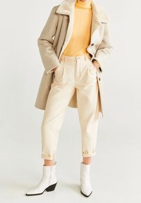 Beige uldfrakke med pelskrave, sennepsfarvet turtleneck, cremede højtaljede bukser og hvide ankelstøvler med spids tå og blokhæl.