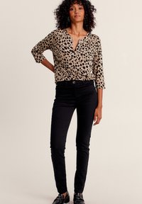 Leopardenmuster-Bluse in Beige und Schwarz, mit V-Ausschnitt und Dreiviertelärmel, kombiniert mit schwarzen, figurbetonten Hosen und schwarzen Schuhen.