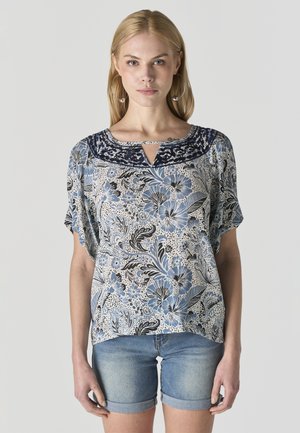 Mujer con una blusa de manga corta azul y blanca con estampado floral y pantalones cortos de mezclilla arremangados, de pie contra un fondo liso.