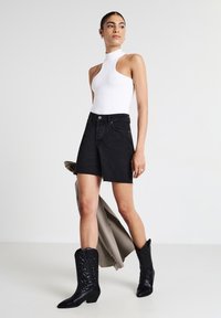 Haut ajusté blanc à col montant, short en denim noir taille haute avec ourlet effiloché, et bottes de cow-boy noires avec des détails gaufrés.
