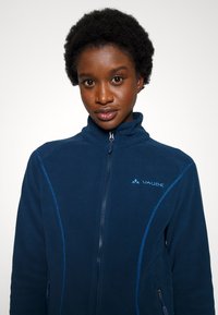 Veste en polaire marine avec col montant, zip intégral, accents de couture bleus et logo sur la poitrine. Texture lisse, conçue pour la chaleur.