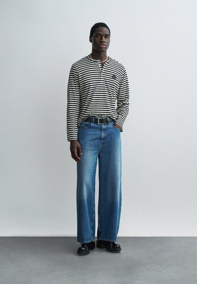 Άνδρας που στέκεται φορώντας μαύρο και άσπρο ριγωτό πουκάμισο μακρυμάνικο, μπλε τζιν παντελόνι με φαρδιά πατζάκια και μαύρη ζώνη, και μαύρα παπούτσια με πλατφόρμα.