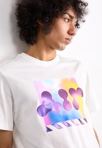 Λευκό βαμβακερό T-shirt με πολυχρωμο γραφικό που περιέχει μωβ σχήματα και κείμενο "Air", σε παστέλ φόντο.